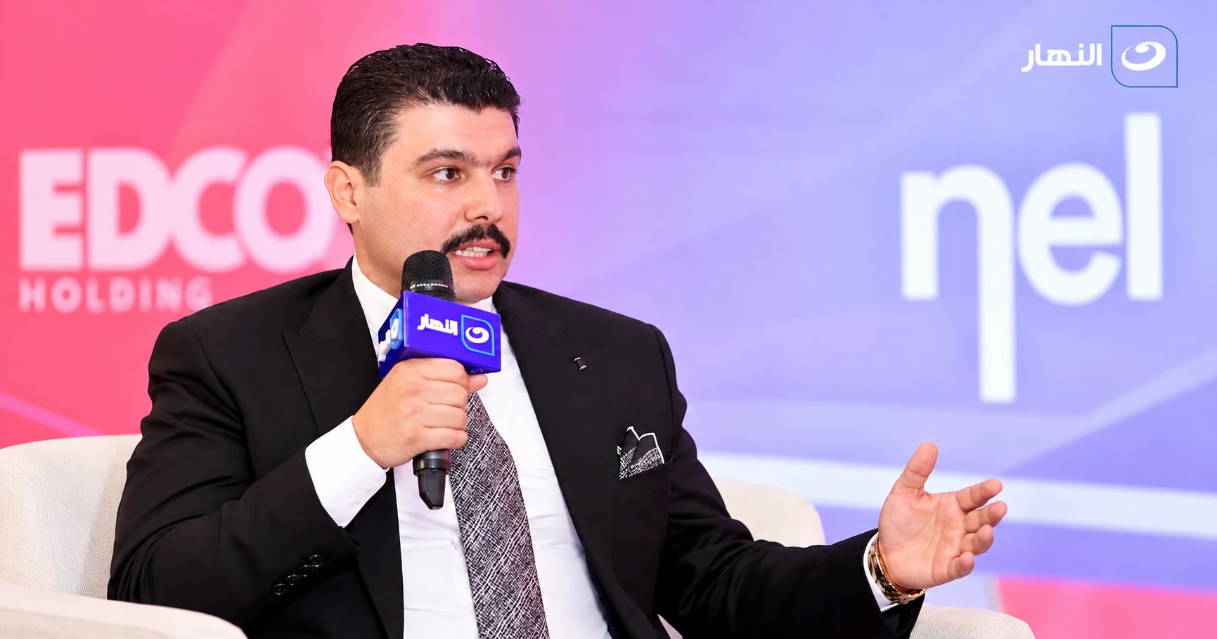 الدكتور المهندس أحمد الزلاط خلال لقاء تلفزيوني يتحدث أمام الكاميرا ممسكًا بميكروفون يحمل شعار قناة النهار